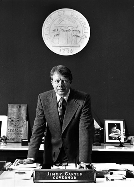 Jimmy Carter standing at a desk with a governor nameplate in the foreground and a State of Georgia seal in the background (black and white image).