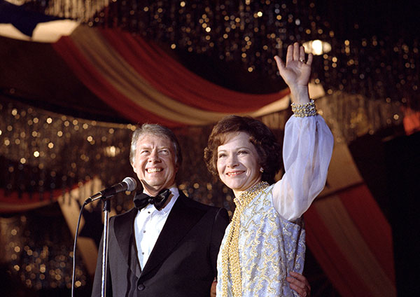 President Carter at a mic with Mrs. Carter as she waves to the crowd.