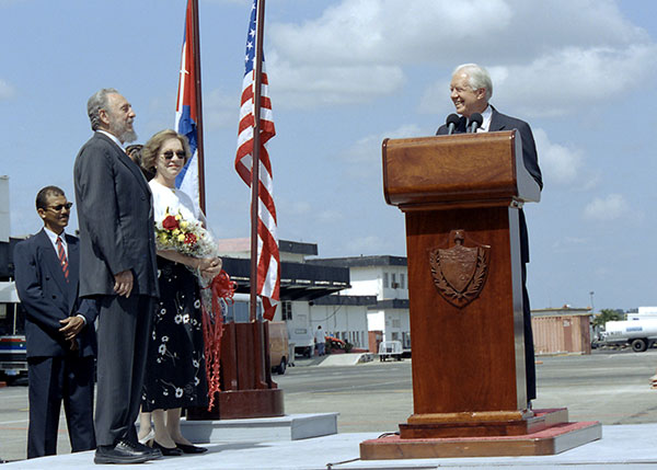 Jimmy Carter at a podium outdoors as Fidel Castro and Rosalynn Carter look on.